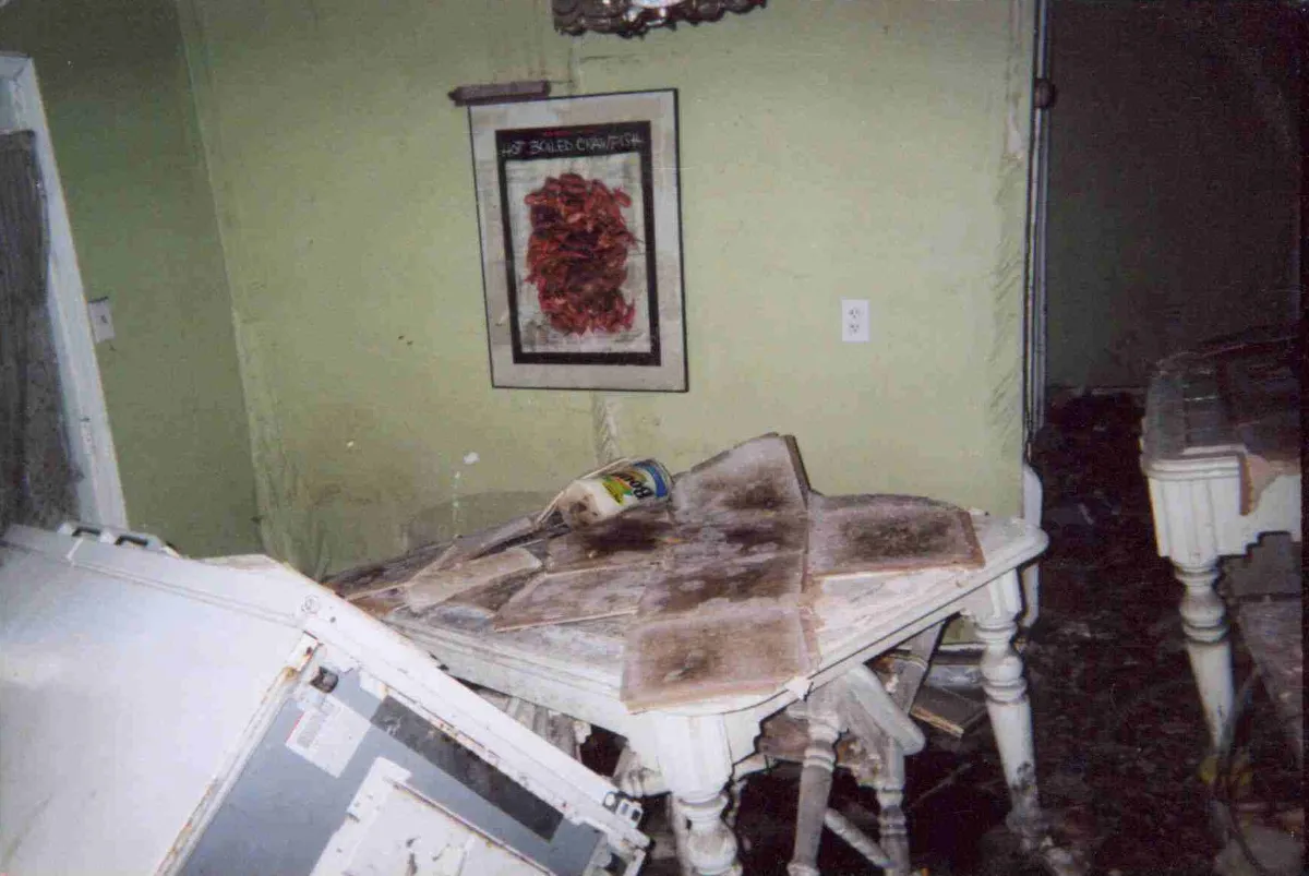 Flood-damaged kitchen after Hurricane Katrina, with a tipped-over refrigerator, fallen ceiling tiles on the table and mud on the floor